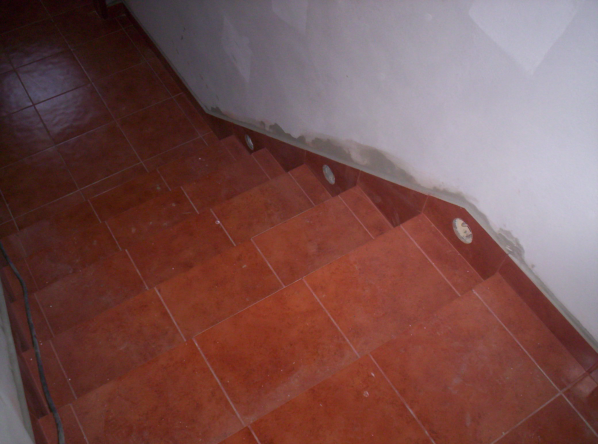 Schody wyłożone pomarańczowo-brązowymi płytkami ceramicznymi z widocznymi otworami na oświetlenie punktowe w ścianie w trakcie remontu.